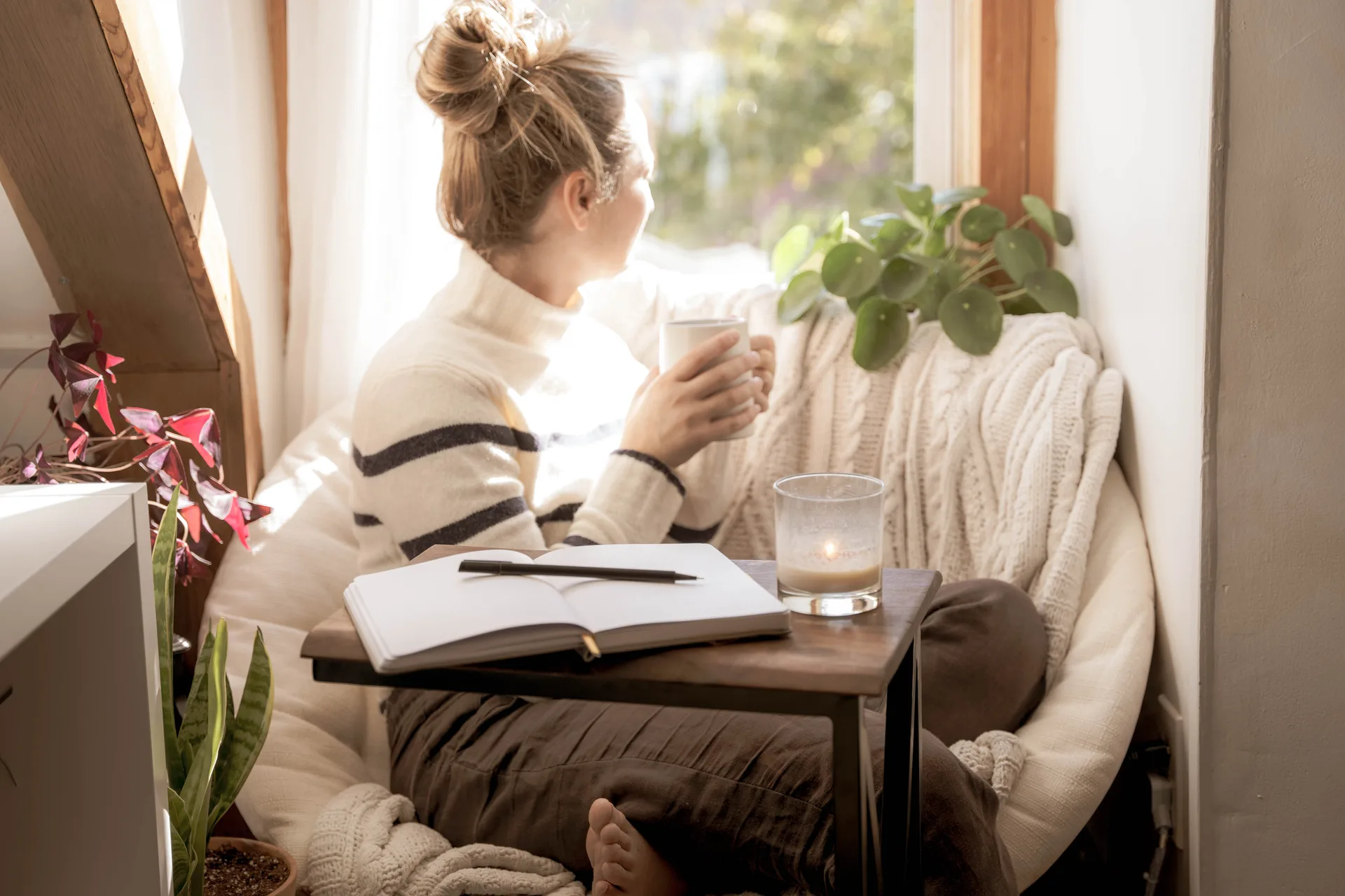 Young woman taking a mental health break to write in her journal stares out the window with coffee in hand
