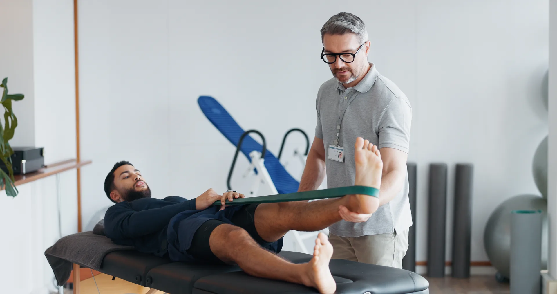 Physiotherapist, people and band for stretching legs at clinic, helping and recovery on table. Men, flexibility exercise and chiropractor for rehabilitation treatment, support and