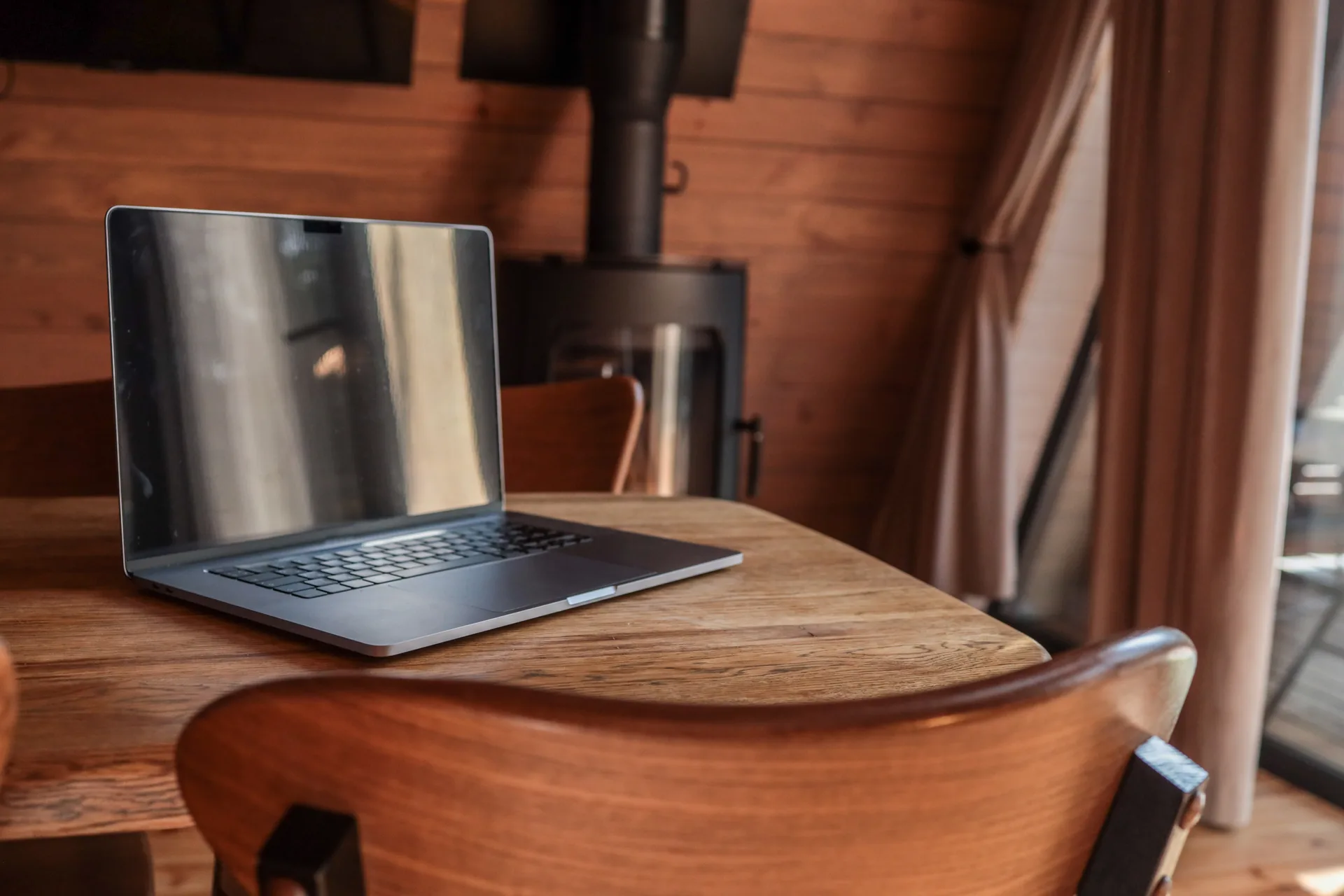 Laptop on a wooden table inside a cozy cabin with a wood-burning stove and warm sunlight streaming through the window, creating a peaceful work-from-home atmosphere